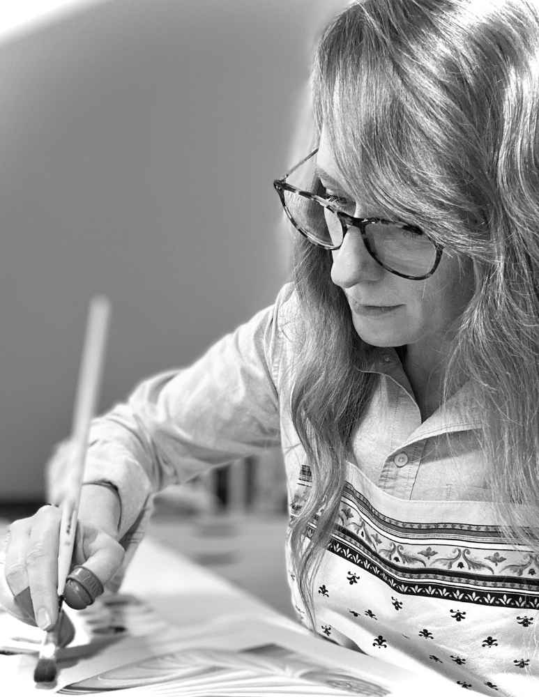 A black and white lifestyle photograph of a the artist at Epiphania Studio with long hair and glasses, wearing a patterned button-up shirt and drawing on paper with a pencil.